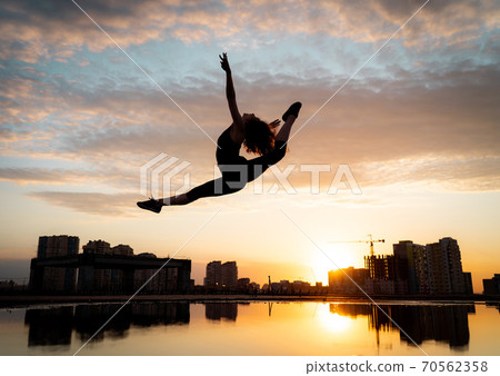 Female flexible dancer jumping during sunset on cityscape background with reflection in the water. Concept of freedom and happiness  70562358