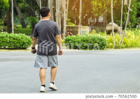 Back view portrait of a Asian man in fitness wear walking and jogging for good health in public park. Jogger in nature. Man enjoying Peaceful nature. Healthcare concept. 70562500