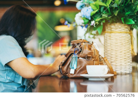 Mini alcohol gel bottle to kill Corona Virus(Covid-19) hang on a brown leather shoulder bag with hot coffee on table in cafe.New normal lifestyle.Health care concept.Selective focus on alcohol gel 70562576