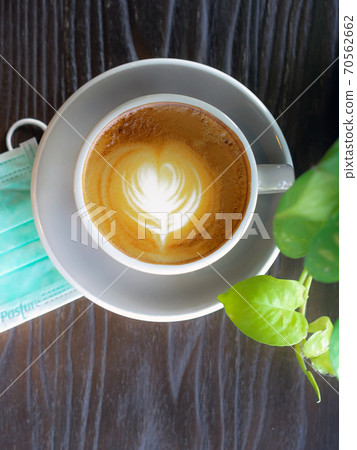 Hot Cappuccino coffee with protective mask on a wooden table for people being quarantined in the situation of the spread of the corona virus (Covid-19).  New normal lifestyle concept. Selective focus 70562662