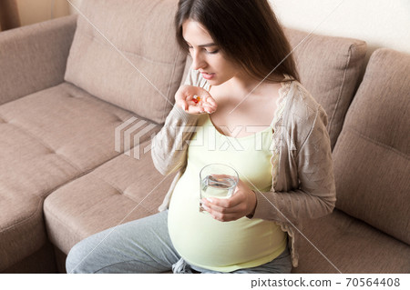 Beautiful woman sitting on sofa holding a pill and water Beautiful woman sitting on sofa holding a pill and water 70564408