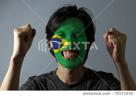 Cheerful portrait of a man with the flag of the Brazil painted on his face on grey background.  70572839