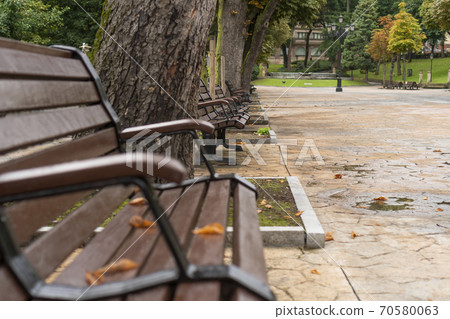 Brown wooden plank bench. varnished, with metal handles in a park with trees. Rest from city bustle Brown wooden plank bench. varnished, with metal handles in a park with trees. Rest from city bustle 70580063
