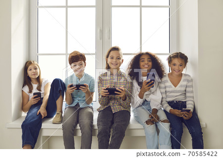 Happy multiethnic children playing games on mobile phones sitting on windowsill Happy multiethnic children playing games on mobile phones sitting on windowsill 70585422