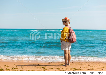 Stylish woman traveler in a summer straw hat with a tourist backpack and a thermo mug back stands and looks at the beautiful seascape 70588954