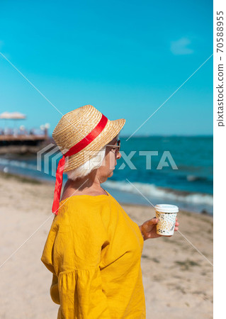 Blonde contented woman holding paper cup with coffee drink while standing by the sea Blonde contented woman holding paper cup with coffee drink while standing by the sea 70588955