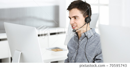 Casual dressed young man using headset and computer while talking with customers online. Call center, business concept Casual dressed young man using headset and computer while talking with customers online. Call center, business concept 70595743