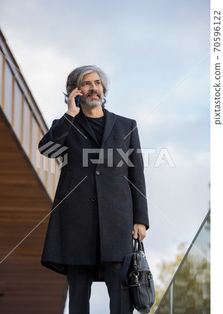 Confident grey-haired businessman in black coat making appointment by phone 70596122