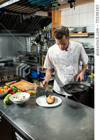 Young male chef in white uniform putting piece of fried salmon on plate Young male chef in white uniform putting piece of fried salmon on plate 70596365