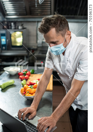 Professional young chef in uniform and protective mask using laptop at work 70596373