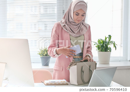 Young pretty Muslim female office manager putting smartphone into her handbag 70596410