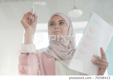 Young serious female broker in hijab holding financial papers and highlighter Young serious female broker in hijab holding financial papers and highlighter 70596413