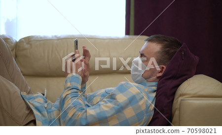 Man in a Medical Mask Uses a Smartphone at Home During Quarantine. Works Alone From Home Remotely. Quarantine Covid-19 Corona Virus. Social Distancing Concept. Man in a Medical Mask Uses a Smartphone at Home During Quarantine. Works Alone From Home Remotely. Quarantine Covid-19 Corona Virus. Social Distancing Concept. 70598021