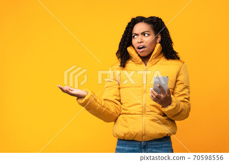 Displeased Black Woman Holding Smartphone Standing On Yellow Studio Background Displeased Black Woman Holding Smartphone Standing On Yellow Studio Background 70598556