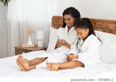 Black mom and little daughter wearing bathrobes chilling in bedroom with smartphones Black mom and little daughter wearing bathrobes chilling in bedroom with smartphones 70598611