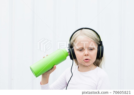 Young blond girl on white background drink fresh spring water from green reusable bottle. Healthy lifestyle in summer city. 70605821