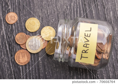 Open tilted glass jar with scattered euro coins with the inscription TRAVEL on a black wooden table. The concept of savings for trips to countries and cities. 70608764