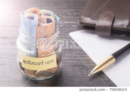 Glass jar with coins and euro banknotes with the words EDUCATION. Old black table. Pen, blank sheet of paper and leather wallet. The concept of savings at the institute, university or college 70609024