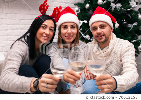 Young happy couple in Christmas hats near a Christmas tree kissing, holding glasses of wine. New Year celebration 70612155