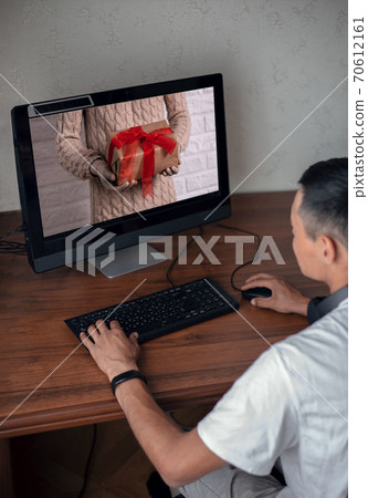Young man sees an image on a computer screen with gloved hands holding a gift. Holiday concept in the context of the coronavirus pandemic. Buying christmas gifts online Young man sees an image on a computer screen with gloved hands holding a gift. Holiday concept in the context of the coronavirus pandemic. Buying christmas gifts online 70612161