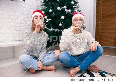 Young happy couple in Christmas hats near a Christmas tree kissing, holding glasses of wine. New Year celebration 70612193