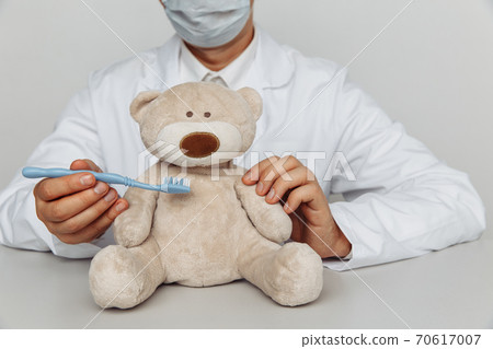 Dentist in mask cleaning teeth of teddy bear at office. Children dentist concept 70617007