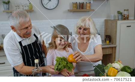 Happy vegan senior couple dancing with granddaughter child while cooking vegetables in kitchen Happy vegan senior couple dancing with granddaughter child while cooking vegetables in kitchen 70617521