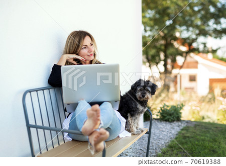 Mature woman with dog working in home office outdoors on bench, using laptop. Mature woman with dog working in home office outdoors on bench, using laptop. 70619388