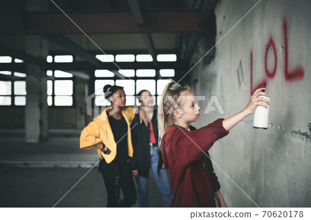 Group of teenagers girl gang indoors in abandoned building, using spray paint on wall. 70620178