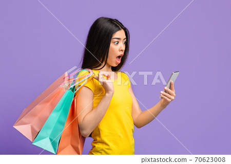 Online Sales. Shocked Asian Woman Looking At Smartphone And Holding Shopping Bags 70623008