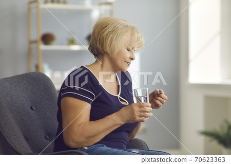 Mature smiling woman sitting in armchair and taking medical pill and pure water in glass in hands 70623363
