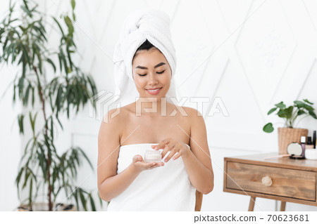 Smiling asian young lady wrapped in towel using cream 70623681