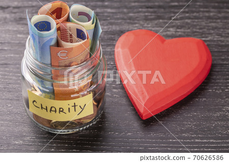 Glass jar with coins and euro notes with the inscription CHARITY. With a huge red heart on an old black wooden table. The concept of kindness and help those in need. Glass jar with coins and euro notes with the inscription CHARITY. With a huge red heart on an old black wooden table. The concept of kindness and help those in need. 70626586