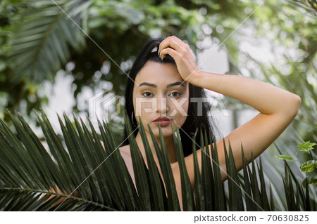 Woman beauty portrait. Sexy attractive Asian model with nude makeup and ponytail hairstyle, touching head and looking at camera, posing against tropical plants. Natural beauty Woman beauty portrait. Sexy attractive Asian model with nude makeup and ponytail hairstyle, touching head and looking at camera, posing against tropical plants. Natural beauty 70630825