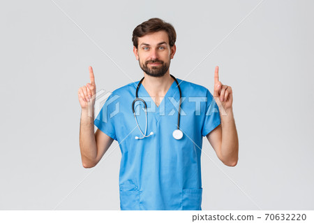 Healthcare workers, pandemic and coronavirus outbreak concept. Handsome bearded doctor in scrubs, stethoscope, pointing fingers up, showing top advertisement, treat patients from covid-19 70632220