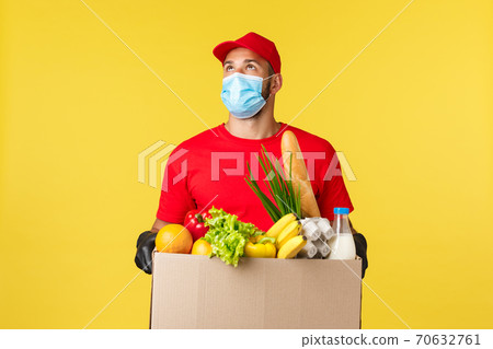 Takeaway delivery, food and drinks, covid-19 quarantine and groceries concept. Courier looking up at building, bring order grocery box to home or office, wearing protective medical mask, gloves 70632761