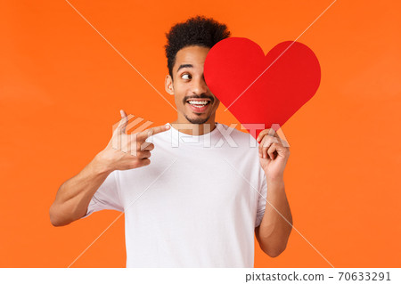 Excited happy, charismatic african-american male in white t-shirt, holding heart valentines card over face, pointing at sign and smiling amused, prepared surprise gift for lover, orange background 70633291