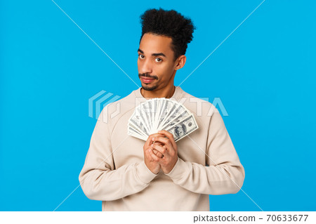 Guy got cash, thinking what buy, how waste money during winter holidays sale season. African american good-looking man with afro haircut holding dollars near chest and gazing suspicious camera 70633677