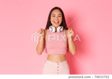 Portrait of cheerful upbeat asian girl in trendy clothes, with headphones around neck, jumping from happiness or anticipation, rejoicing over great news, winning something, pink background 70633702