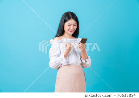 Asian woman holding smart phone with fist hand and excited for success on light blue background. Asian woman holding smart phone with fist hand and excited for success on light blue background. 70635854