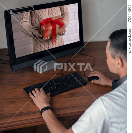 Young man sees an image on a computer screen with gloved hands holding a gift. Holiday concept in the context of the coronavirus pandemic. Buying christmas gifts online Young man sees an image on a computer screen with gloved hands holding a gift. Holiday concept in the context of the coronavirus pandemic. Buying christmas gifts online 70644805
