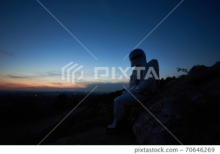 Astronaut sitting on rocky hill during sunset. Astronaut sitting on rocky hill during sunset. 70646269