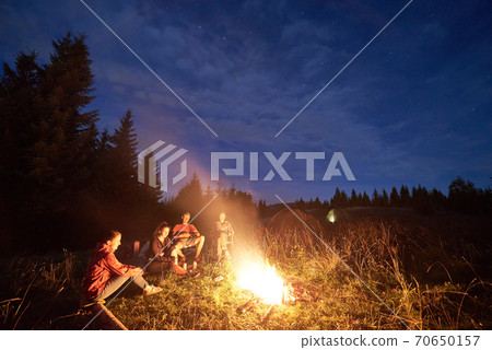 Group of friends sitting around a bonfire on a green mountain meadow surrounded by spruce trees under starry sky Group of friends sitting around a bonfire on a green mountain meadow surrounded by spruce trees under starry sky 70650157