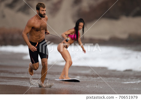 A young man pulls a young girl on a rope who is learning a skimboard 70651979