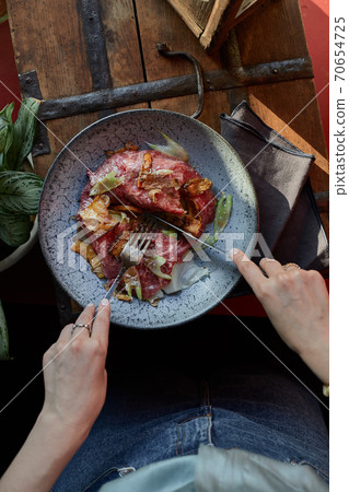 A female hand is preparing a vegetable salad with beef roast beef. A plate of salad with beef roast beef, onions, arugula, pepper and tomatoes on a black table. Top view. 70654725