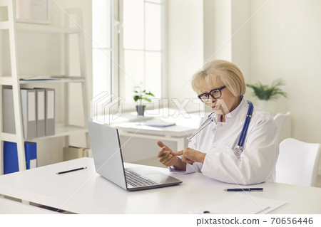 Mature woman doctor in uniform and glasses consulting patient online on laptop 70656446