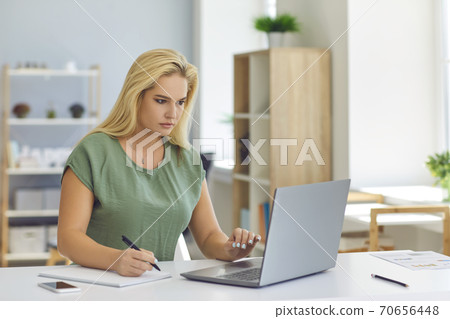 Woman sitting at desk, working online with laptop and making notes Woman sitting at desk, working online with laptop and making notes 70656448