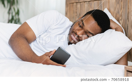 African american guy using cellphone lying in bed in bedroom 70658918