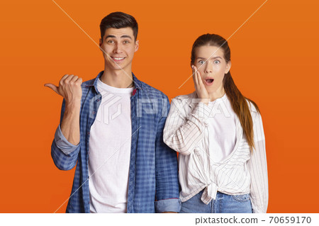 Surprised man smiles and points finger to free space, amazed female with open mouth presses hand to her cheek 70659170