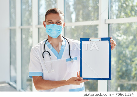 Confident male doctor wearing medical mask showing clipboard with diagnosis in hospital background. 70661154